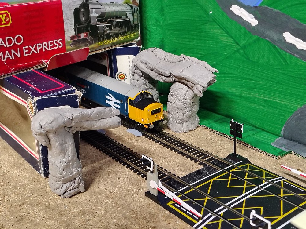 A blue Class 37 model emerging from a hand-made tunnel mouth on sectional track, with train-set boxes stacked behind the scenery.