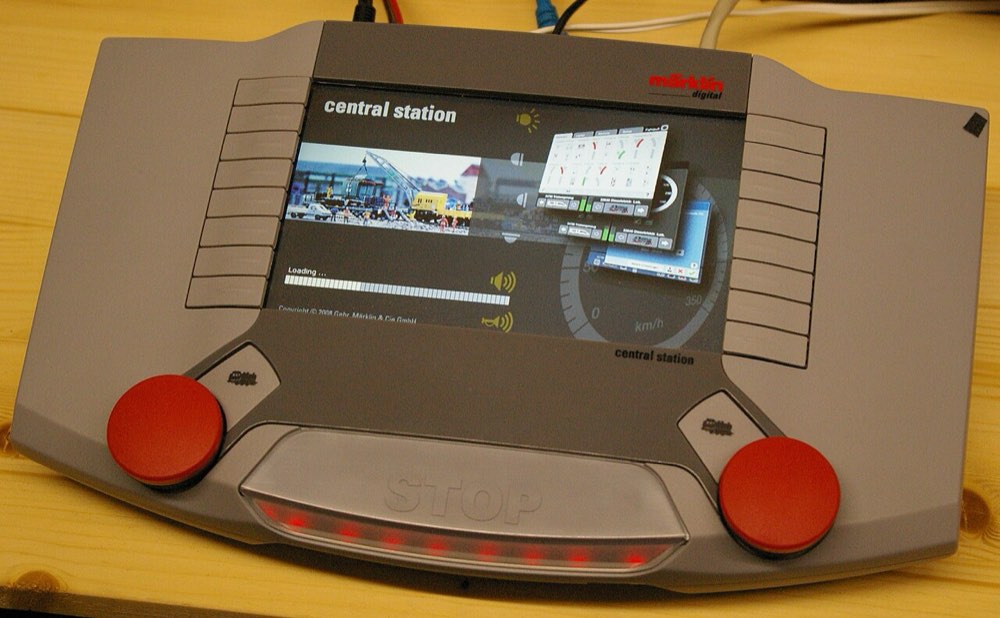 A Marklin Central Station 2 command station: a grey wedge-shaped unit with a colour touchscreen, two red control knobs and a lit STOP bar.