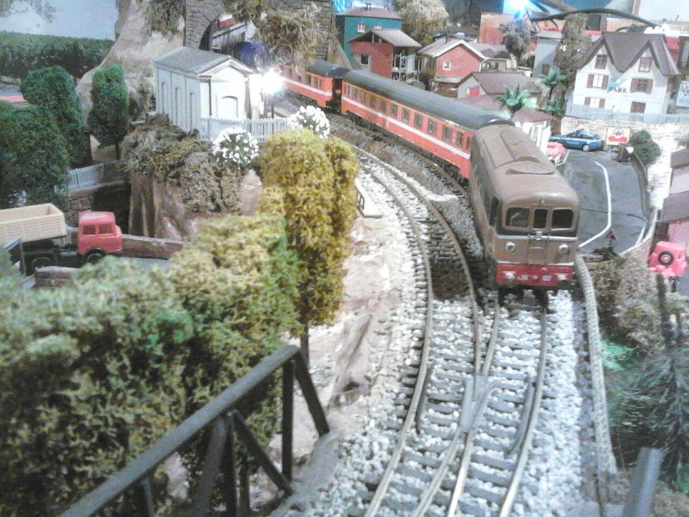 An Italian-outline diesel and red coaches curving through a model town, with ballasted track and a turnout in the foreground.
