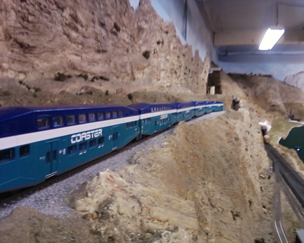 An HO scale model of a Coaster bi-level commuter train running through a rocky cutting on a large basement layout.