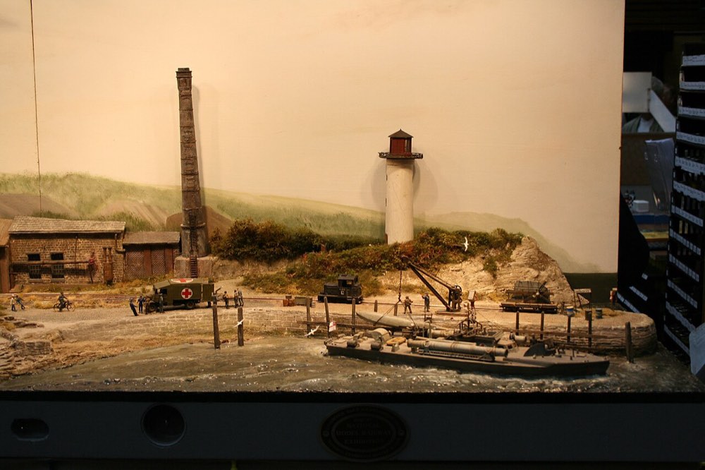 A wider view of the 009 quayside layout, showing a lighthouse, a tall brick chimney, quayside buildings and a painted backdrop behind.