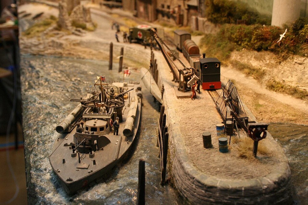 A 009 narrow-gauge quayside layout: a gun boat alongside the wall, a small industrial tank engine and wagons on the cobbles, and a crane at work.