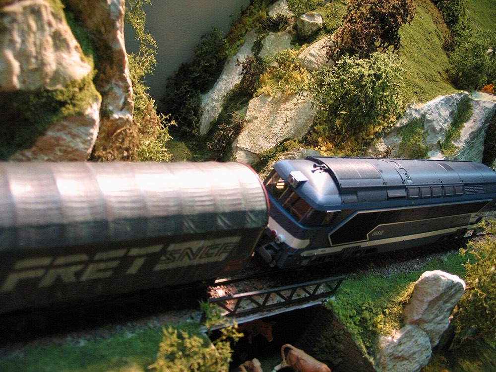 A model SNCF diesel locomotive hauling a covered freight wagon across a small girder bridge through rocky, mossy scenery.