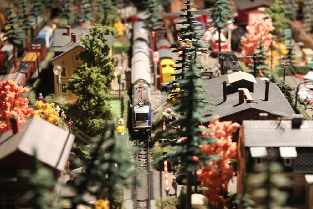 A model town seen from above: a modern multiple unit threading between houses, conifers and autumn-coloured trees at a level crossing.